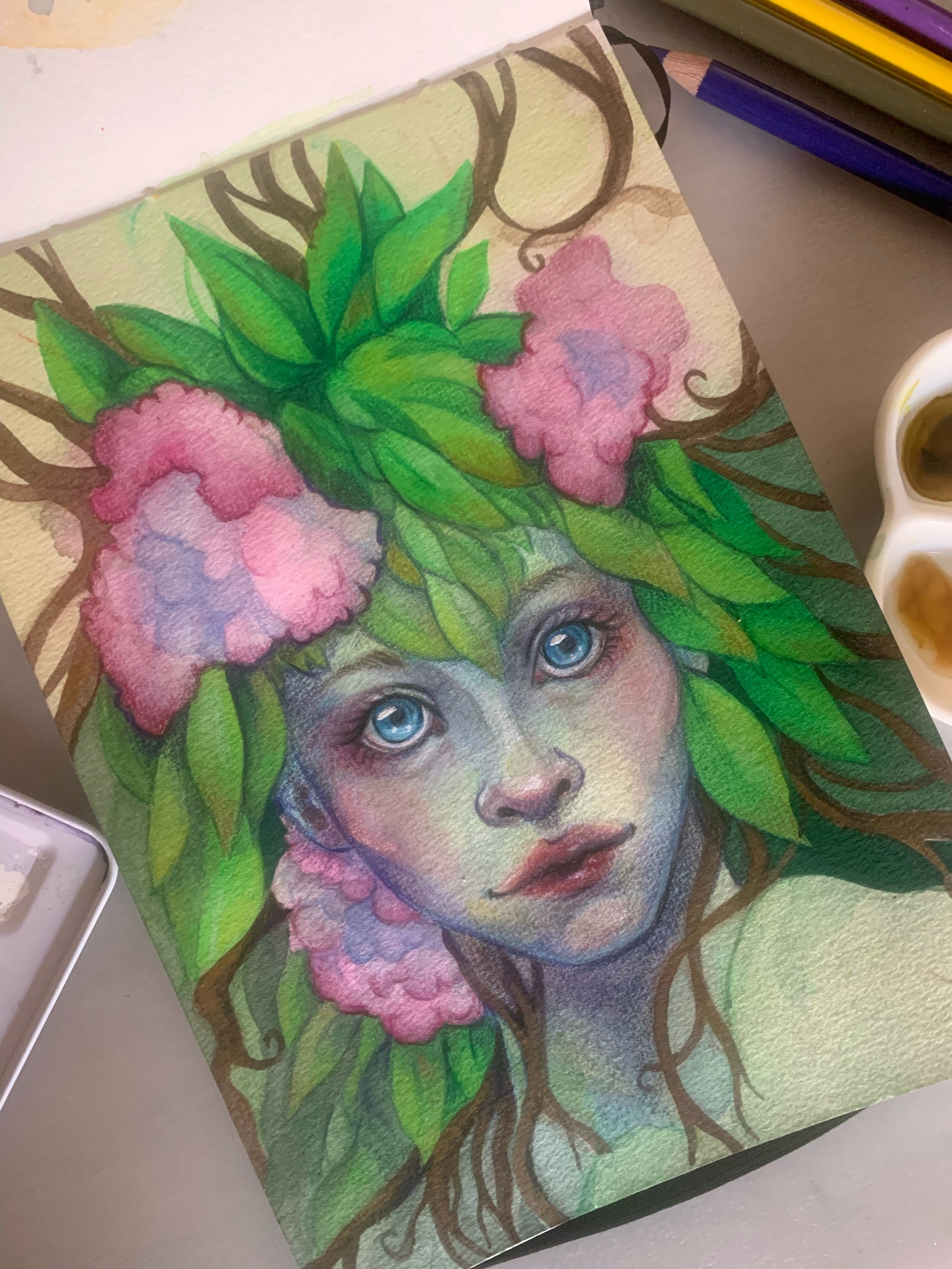 Portrait d'une femme avec une chevelure de feuilles et de fleurs. Ses yeux sont bleus. Autour, on retrouve du matériel de dessin (des pots de peintures, des crayons de couleurs et des pinceaux)